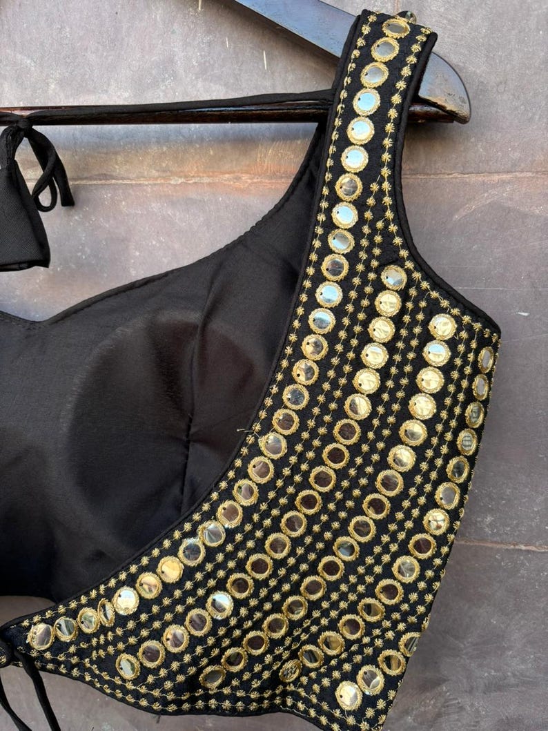 May include: A black sleeveless blouse featuring a wide shoulder strap adorned with multiple rows of circular reflective mirrors and gold-toned embroidery. The garment has a sweetheart neckline and includes thin black fabric ties at the shoulder.
