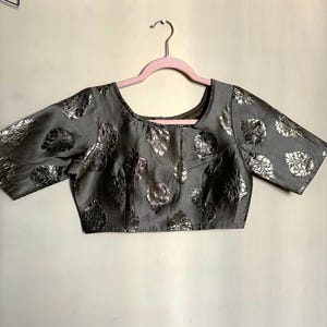 May include: A gray blouse with silver floral patterns. The short-sleeved top has a square neckline and is hanging on a pink hanger. The fabric has a subtle sheen, and the floral design is repeated across the garment.