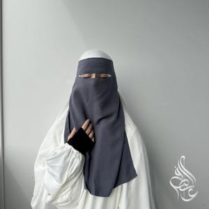 Puede incluir: Un niqab gris oscuro con un cubrecabezas a juego y un vestido interior blanco. El niqab cubre la parte inferior de la cara, con una abertura para los ojos. Las mangas son blancas con puños negros. El fondo es una pared gris claro lisa.
