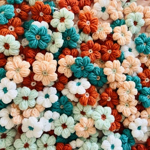May include: A collection of crocheted flowers in various colors, including peach, white, teal, and orange. The flowers are arranged in a random pattern and are all approximately the same size.