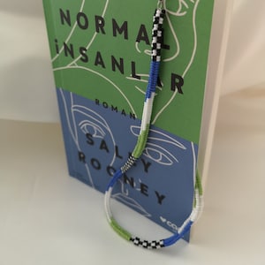 May include: A colorful beaded bookmark with a silver clasp is attached to a book titled "Normal Insanlar" by Sally Rooney. The bookmark features a pattern of black, white, blue, and green beads. The book has a green cover with white line art.