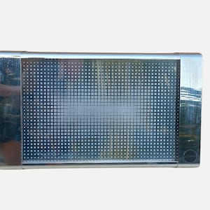 Può includere: Un pannello luminoso rettangolare con una cornice in cromo lucido e uno schermo centrale con una fitta griglia di piccoli punti bianchi che aumentano di dimensioni verso il centro, creando un effetto sfumato. Il dispositivo ha un tasto circolare.