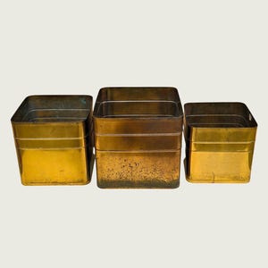 Könnte beinhalten: Ein Set aus drei quadratischen Messingbehältern in unterschiedlichen Höhen, die jeweils eine glatte metallische Oberfläche mit sichtbaren Anzeichen von Oxidation und Patina aufweisen. Die Behälter haben gerade Seiten und offene Oberseiten.
