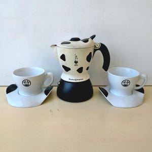 Puede incluir: Una cafetera de estufa Bialetti Mukka Express con un patrón de manchas de vaca en blanco y negro, con base y asa negras. Se incluyen dos tazas y platillos de cerámica blanca a juego, cada uno con un pequeño icono de cara de vaca negra.