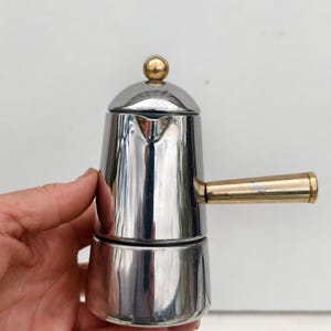 May include: A stainless steel stovetop espresso maker with a gold-colored handle and top knob. The coffee pot is cylindrical with a two-part design. The handle is attached to the side, and the top knob is a small sphere.