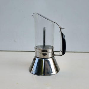 May include: A stovetop espresso maker featuring a clear glass upper chamber with a central metal rod, a polished stainless steel base, and a black plastic handle attached to the side. The design includes a hinged metal lid and a spout.