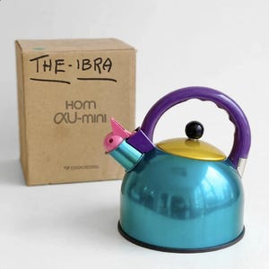Può includere: Un bollitore da tè in metallo color verde acqua con manico viola, coperchio giallo, pomello nero e beccuccio rosa. Dietro si trova una scatola di cartone con la scritta THE-IBRA HOM XU-mini COOKVESSEL. Il bollitore ha un corpo arrotondato.