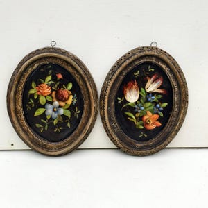 以下が含まれることがあります： 2つの楕円形の花の絵。各絵画は、バラやチューリップなどのカラフルな花が描かれた暗い背景が特徴です。絵画は、装飾的で質感のある金色のフレームに収められています。