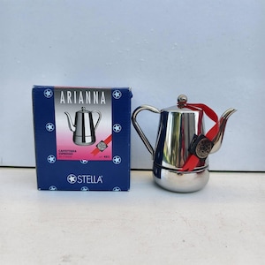 Peut inclure: Une cafetière italienne en acier inoxydable brillant avec une poignée et un bec courbés, à côté d'une boîte bleue avec le texte "ARIANNA" et une image de la cafetière. La cafetière a un ruban rouge et un sceau noir.