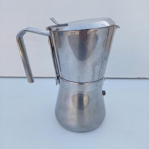May include: A stainless steel stovetop espresso maker with a handle and lid. The coffee pot has a classic design, with a cylindrical base and a tapered upper chamber. The handle is a sturdy, curved metal. The lid has a small, hinged flap.