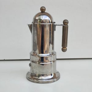 Puede incluir: Una cafetera italiana de acero inoxidable con tapa redondeada y asa. La cafetera tiene una superficie brillante y reflectante y un pequeño pico. El asa es oscura y cilíndrica.