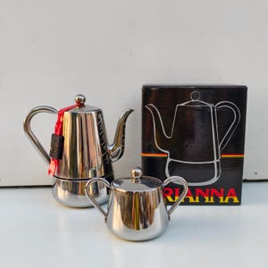 Könnte beinhalten: Ein Edelstahl-Kaffeezubereiter und Sahne-Set. Der Kaffeezubereiter hat einen langen Ausguss und einen gebogenen Griff. Der Sahnebereiter hat zwei Griffe und einen Deckel. Eine schwarze Box mit dem Namen "RIANNA" ist ebenfalls im Bild.