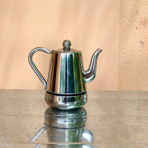 May include: A shiny, stainless steel stovetop espresso maker with a rounded base, a cylindrical body, and a curved handle. The coffee pot has a spout and a small, round knob on the lid. The pot is sitting on a reflective surface.