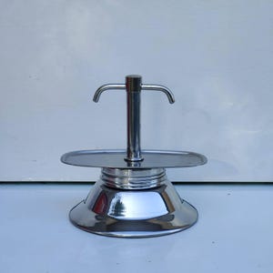 Op de afbeelding: Een zilveren metalen koffiezetapparaat met een ronde basis en een tuit aan de bovenkant. Het koffiezetapparaat heeft een handvat aan de zijkant.