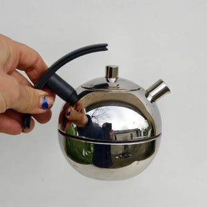 Peut inclure: Une théière sphérique en acier inoxydable avec une poignée et un bec noirs. La théière a une surface brillante et réfléchissante. La poignée est incurvée et fixée au sommet de la théière. Le bec est court et incliné.
