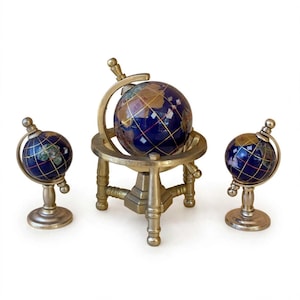 Peut inclure: Un ensemble de trois globes composés de sphères en pierre bleu foncé avec des lignes de grille dorées incrustées et des continents aux tons terreux. Chaque globe est monté sur un support métallique doré avec une base circulaire et un axe.