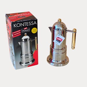 May include: A polished stainless steel Kontessa espresso maker by Vev Vigano with a gold-toned handle and lid knob. The coffee maker is displayed next to its original black and red cardboard box featuring the product name and brand logo.