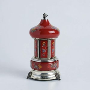 May include: A red carousel-style music box featuring hand-painted floral motifs in yellow, blue, and green. The structure includes a silver-toned base with three ornate feet, a central rotating section with vertical panels, and a domed top with a small cherub.