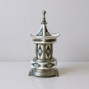May include: A white porcelain pagoda-shaped music box featuring intricate blue and gold floral patterns, metallic silver-toned accents, a tiered roof structure, and a decorative base with ornate feet, topped with a small silver-toned finial.