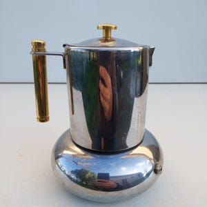 May include: A stainless steel stovetop espresso maker with a gold-colored handle and lid knob. The coffee pot has a round base and a cylindrical body, reflecting the surrounding environment. A classic kitchen appliance.