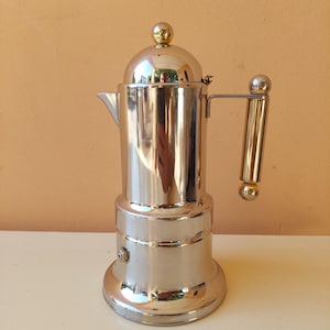 Pode incluir: Uma cafeteira de metal prateado polido para fogão com corpo cilíndrico, tampa abobadada com uma pequena esfera dourada no topo e uma alça vertical acentuada com esferas douradas na parte superior e inferior, com uma pequena válvula lateral.