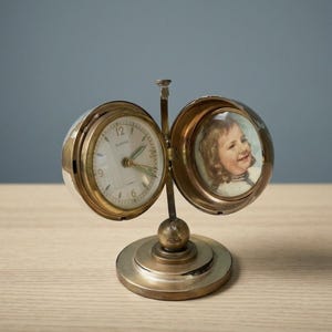 May include: A brass Europa travel clock with a hinged design that opens to reveal a clock face on one side and a circular portrait of a child on the other. The clock features a round base, a central vertical support, and a white dial with numerals.