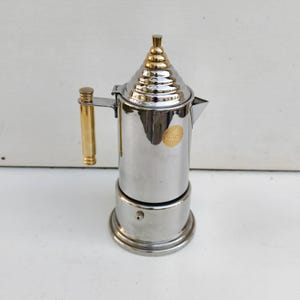 Pode incluir: Uma cafeteira de espresso de aço prateado e dourado com uma pega dourada e uma tampa dourada. A tampa tem um design empilhado com uma parte superior arredondada.