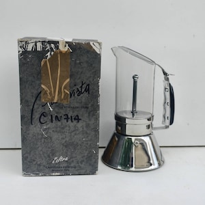 Pode incluir: Uma cafeteira espresso Cinzia com câmara superior de vidro transparente, base metálica e pega lateral, ao lado da sua caixa de cartão cinzenta original com o texto Cinzia, La Vetrina del Caffe e Made in Italy impresso na frente.