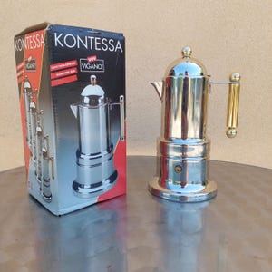 May include: A polished stainless steel Kontessa espresso maker by Vev Vigano with a gold-tone handle and lid knob. The coffee maker is displayed next to its original black and red cardboard box, which features the product name and brand logo.