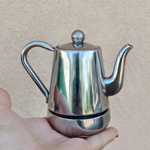 May include: A stainless steel stovetop espresso maker with a curved handle and spout. The small coffee pot has a round lid with a small knob. The shiny metal reflects the surrounding environment. The espresso maker is held in a hand.