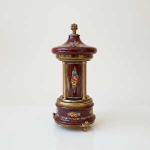 May include: A deep burgundy ceramic music box with a glossy finish, featuring ornate brass-toned metal accents and a central column. The piece is decorated with colorful floral motifs in red, blue, and yellow, topped with a small brass finial.