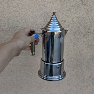 Könnte beinhalten: Ein polierter Espressokocher aus Edelstahl mit zylindrischem Körper, gestuftem konischem Deckel und vertikalem Griff. Die metallische Oberfläche reflektiert das Licht und ein kleines kreisförmiges Emblem ist an der oberen Seite zu sehen.