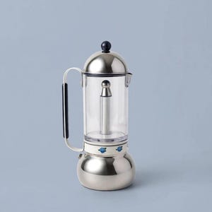 Puede incluir: Una cafetera de estufa con base y tapa de acero inoxidable pulido, cámara cilíndrica de vidrio transparente y mango negro. La sección central presenta una banda blanca decorada con pequeños motivos florales azules y un vástago central.