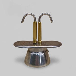 May include: A silver-toned metal stovetop espresso maker featuring a wide, oval-shaped top platform with two circular indentations. Two vertical gold-toned cylindrical handles extend upward, each topped with a curved silver spout facing outward.