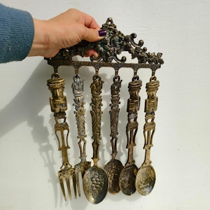 May include: A set of six antique-style brass spoons and a fork hanging on a decorative wall mount. The spoons and fork have ornate handles with a stylized human figure design.