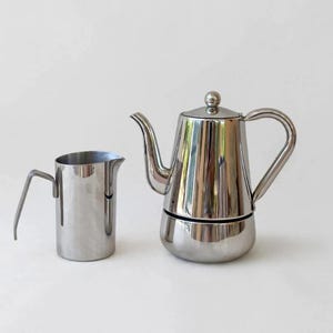 Puede incluir: Una cafetera de acero inoxidable pulido con cuerpo cónico, pico curvo y asa a juego, junto a una jarra de leche cilíndrica de acero inoxidable con un asa angular y un pequeño pico vertedor.