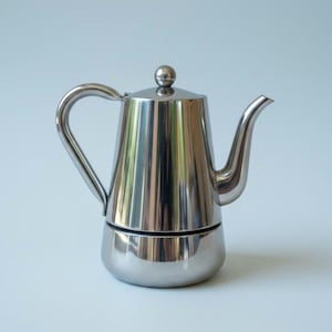 Puede incluir: Una cafetera de acero inoxidable pulido con cuerpo cilíndrico cónico, un asa curva en el lado izquierdo, un pico vertedor delgado a la derecha y un pequeño pomo esférico centrado en la parte superior de la tapa extraíble.