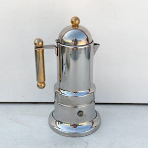 May include: A stainless steel stovetop espresso maker with a gold-colored handle and top. The coffee pot has a shiny, reflective surface and a rounded base. The handle and top are a contrasting gold color.