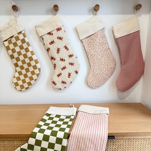 May include: A selection of Christmas stockings with diverse patterns: brown and white check, gingerbread men, red and white stripes, and green and white check. Each stocking features a cream cuff and hangs from a wooden peg.
