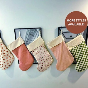 May include: A selection of Christmas stockings in different designs and hues. The stockings feature a solid pink, a red and white floral print, a pink and white striped, and a green and white checkered pattern. Each stocking has a cream-coloured cuff. The image includes the text "MORE STYLES AVAILABLE!"
