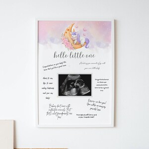 Puede incluir: Una impresión enmarcada en blanco con un fondo de acuarela rosa y morado que presenta un unicornio durmiendo en una luna creciente. La impresión tiene el texto "hello little one" e incluye una imagen de ultrasonido en blanco y negro.