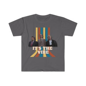 May include: A dark grey t-shirt with a retro-style design featuring three men in suits standing in front of a colourful striped background. The text "IT'S THE VIBE" is printed in yellow and orange letters.