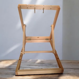 Può includere: Un supporto in legno per telaio da ricamo, con un design pieghevole. Il supporto è costruito in legno di colore chiaro e ha un telaio rettangolare con due ganci metallici. Il supporto è progettato per contenere un telaio da ricamo.