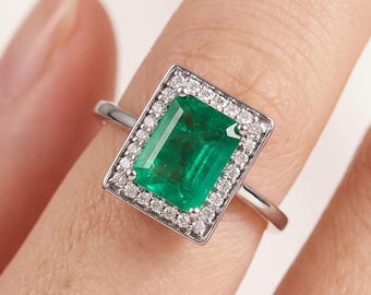May include: A silver ring featuring a rectangular emerald-cut green gemstone, surrounded by a halo of smaller, clear gemstones. The ring is set on a slender silver band, showcasing the vibrant green of the central stone.