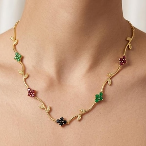 Puede incluir: Un collar de tono dorado con una delicada cadena en forma de enredadera con pequeñas hojas y cinco dijes de flores de cuatro pétalos. Las flores están engastadas con pequeñas piedras redondas en colores alternos de verde, rojo y negro.