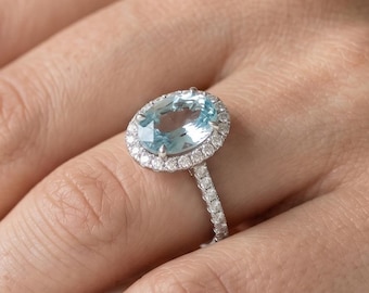 May include: A silver ring with a large, oval-shaped, light blue gemstone, surrounded by a halo of smaller, clear stones. The ring's band is also encrusted with small, clear stones. The ring is on a person's finger.