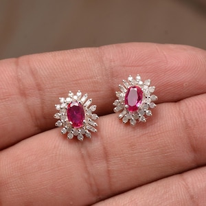 May include: A pair of ruby stud earrings. Each earring has an oval, deep red ruby at the centre, encircled by a halo of small, clear accent stones. The earrings are set in a silver-toned metal.