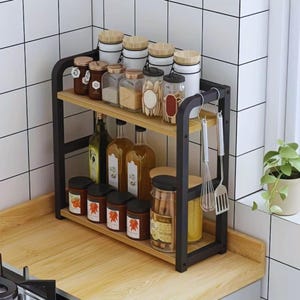 Puede incluir: Un estante de cocina de dos niveles con estructura de metal negro y estantes de madera clara. Contiene varios frascos, botellas y recipientes de vidrio llenos de especias y aceites. Utensilios cuelgan del lateral y una pequeña planta está cerca.
