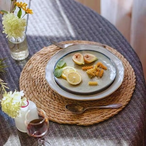 Può includere: Una scena culinaria rustica con un sottobicchiere intrecciato, un piatto con fichi, pasta e fette di limone e un bicchiere di vino rosso. Sono visibili anche un piccolo vaso con fiori gialli e un cucchiaio.