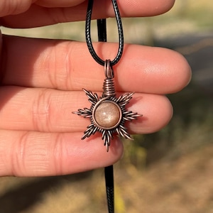 May include: A sun-shaped pendant featuring a round, shimmering tan gemstone encased in intricate copper wire wrapping. The pendant is suspended from a simple black cord, with the wire forming radiating points around the central stone.